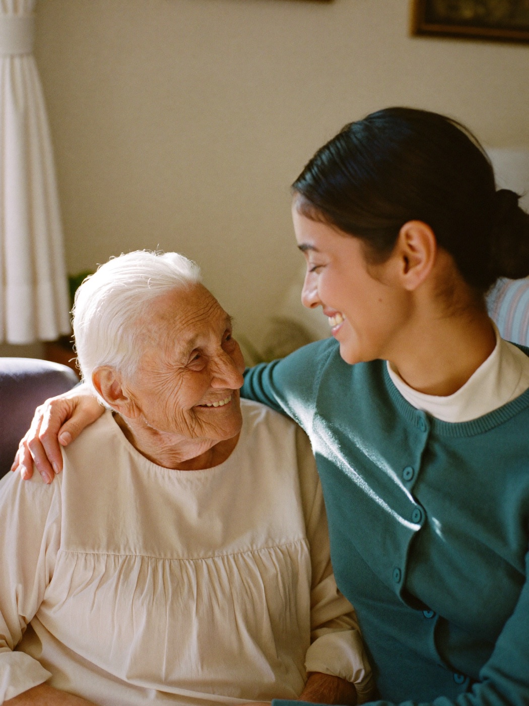 An elderly woman smiling warmly at home while her adult family caregiver rests a hand on her shoulder